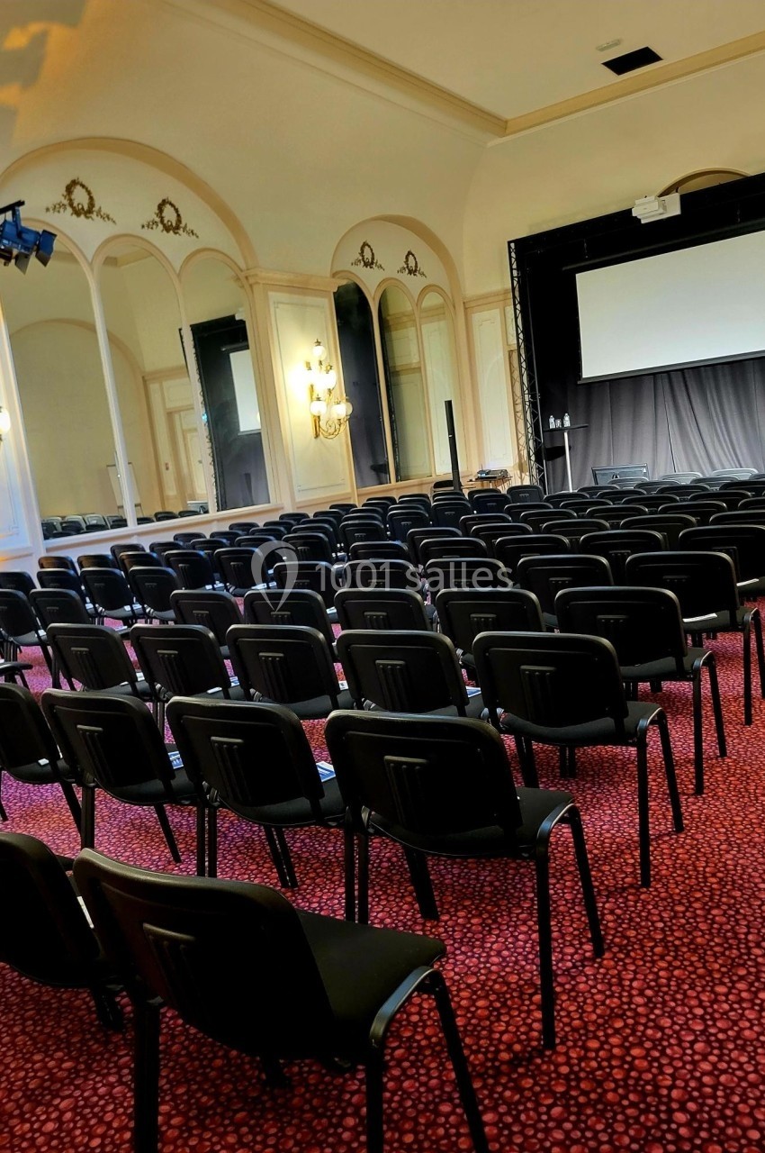 Salle de conférence avec rangées de chaises noires alignées, écran de projection et miroirs sur les murs.
