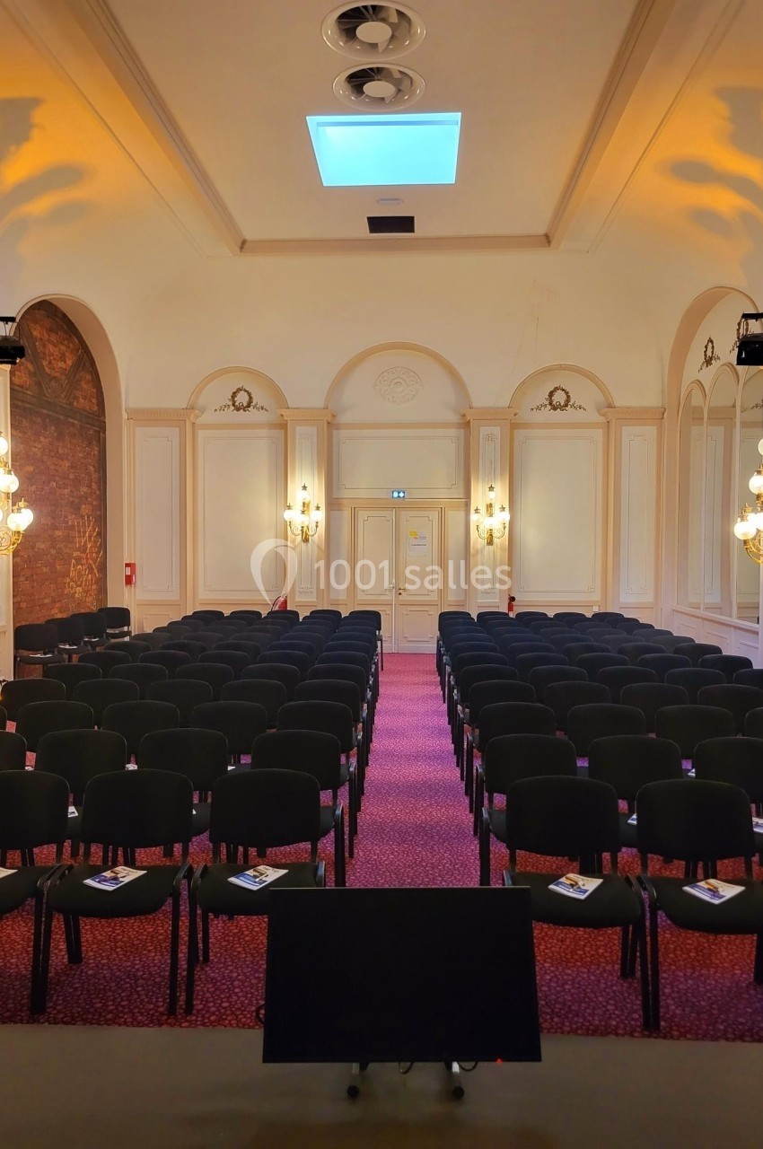 Salle de conférence avec rangées de chaises noires alignées, éclairage chaleureux et écran au premier plan.