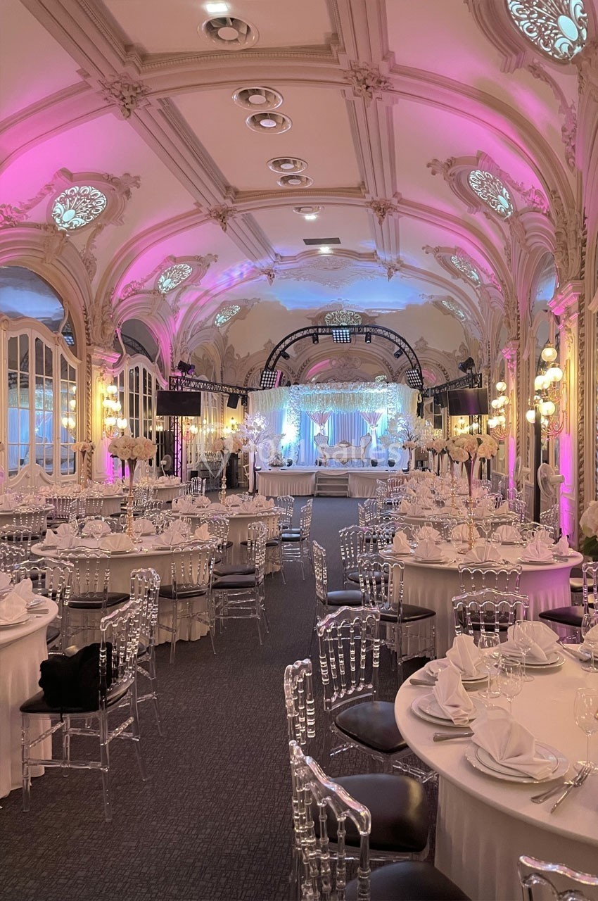 Salle de réception élégante avec tables rondes dressées, chaises transparentes et éclairage rose et blanc.