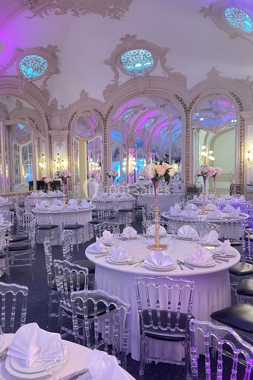 Salle de réception élégante avec tables rondes dressées, chaises transparentes et décorations florales sous un éclairage…