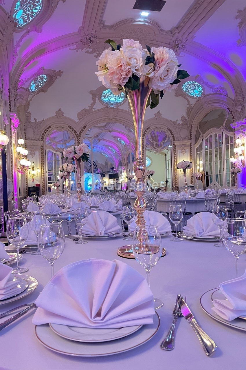 Salle de réception élégante avec tables dressées, nappes blanches, serviettes pliées et centres de table fleuris.