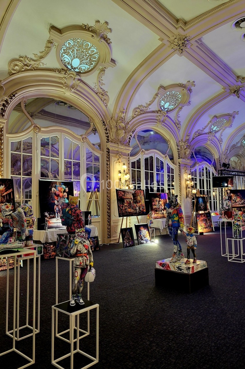 Salle ornée de moulures et vitraux, exposant des sculptures colorées sur des socles et des tableaux lumineux.