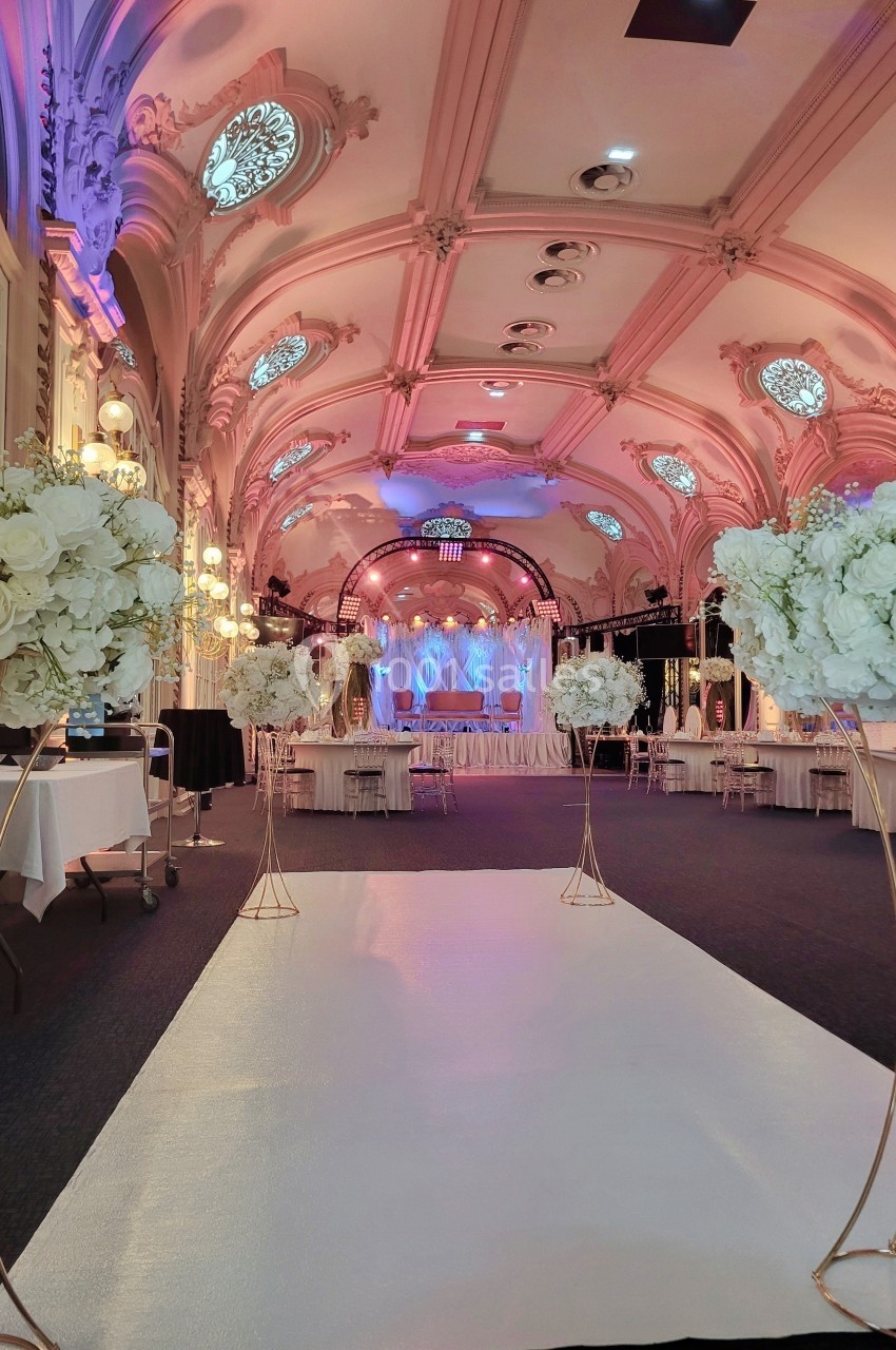 Salle de réception élégante avec plafond orné, allée blanche et décorations florales blanches sur supports dorés.