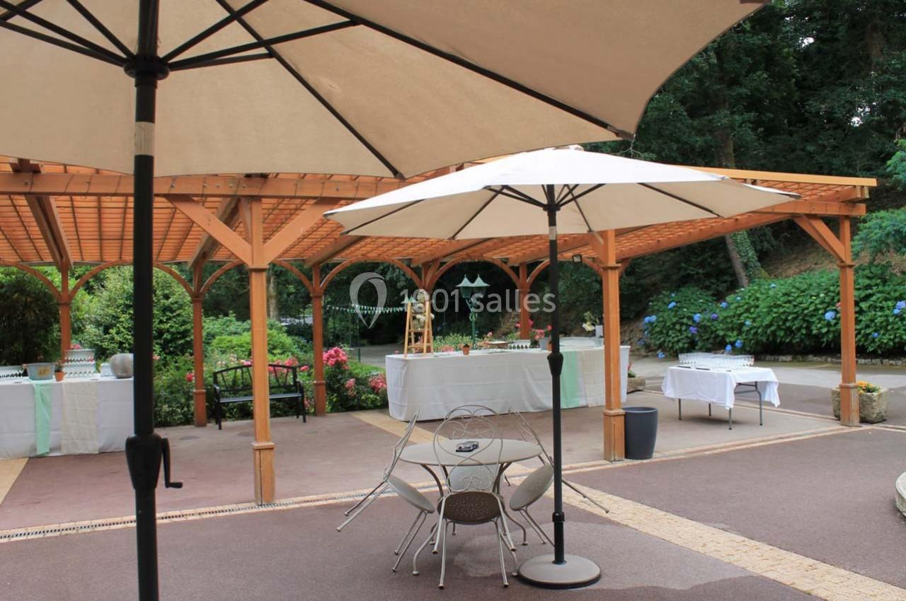Terrasse extérieure avec pergolas en bois, tables, chaises et parasols, entourée de végétation et de fleurs.