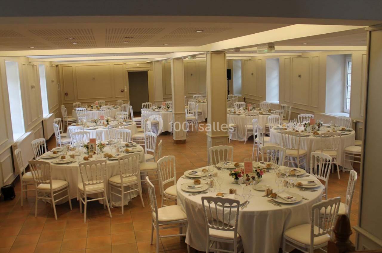 Salle de réception lumineuse avec des tables rondes dressées, décorées de fleurs et entourées de chaises blanches.