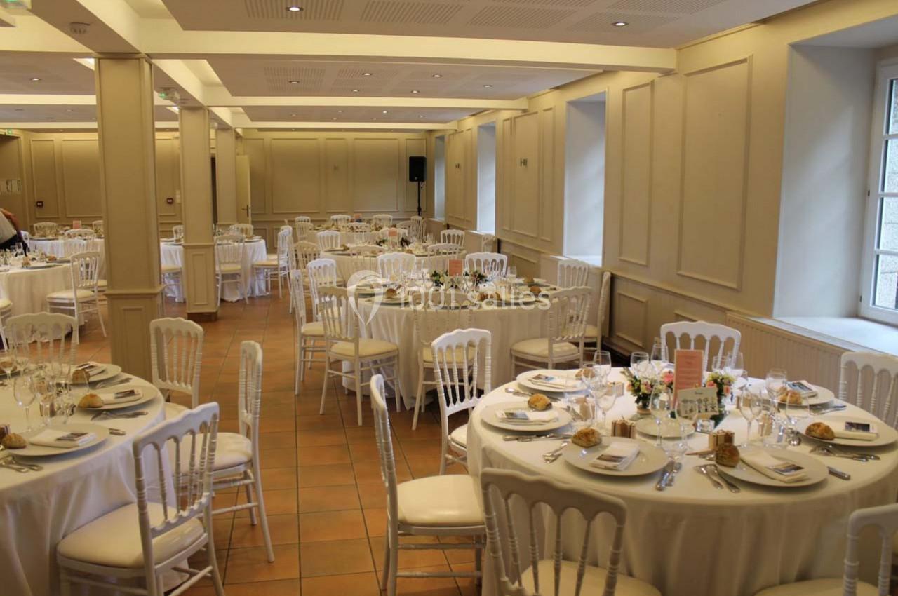 Salle de réception lumineuse avec des tables rondes dressées pour un repas, décorées de nappes blanches et de fleurs.