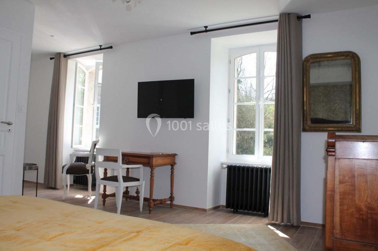 Chambre lumineuse avec lit, bureau en bois, télévision murale, fenêtres avec rideaux et radiateur noir.