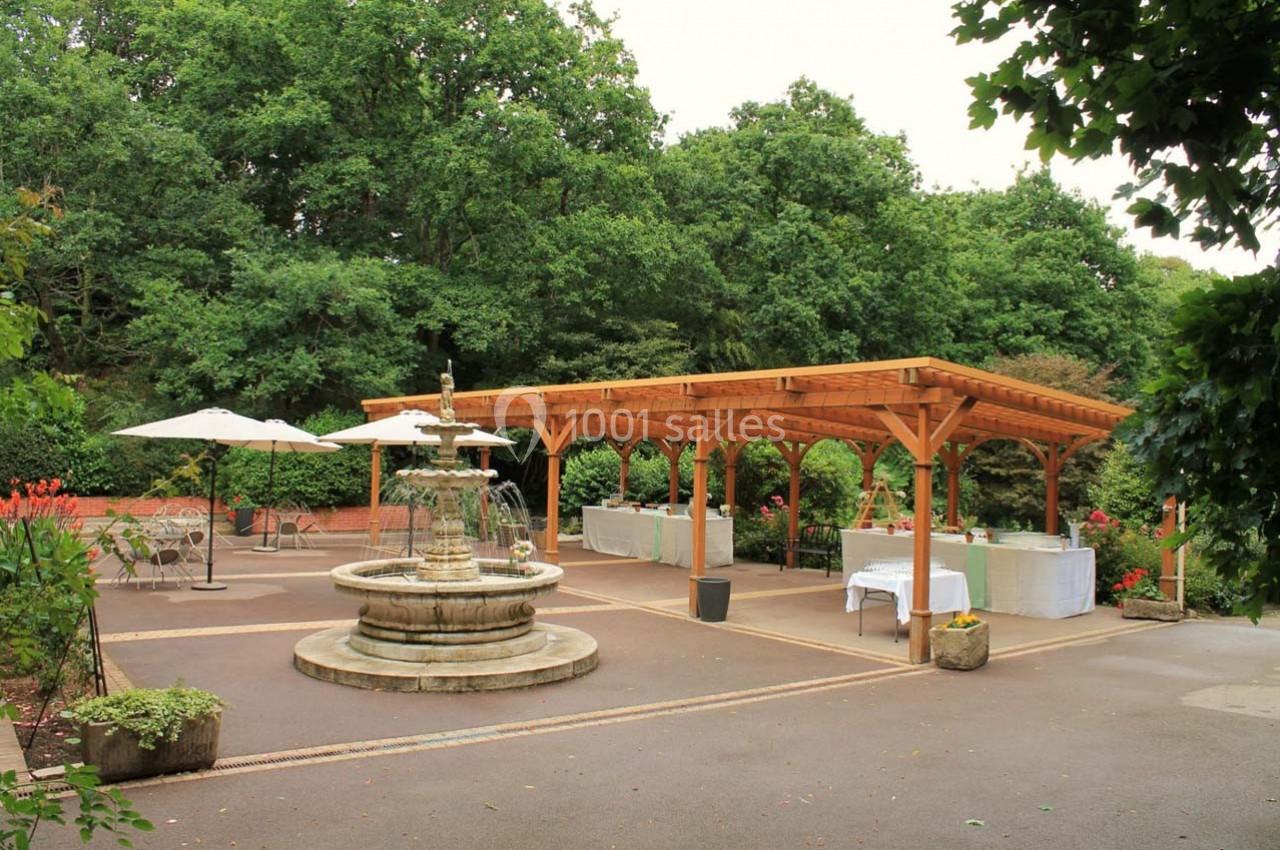 Espace extérieur avec fontaine centrale, pergola en bois, tables dressées et végétation environnante.