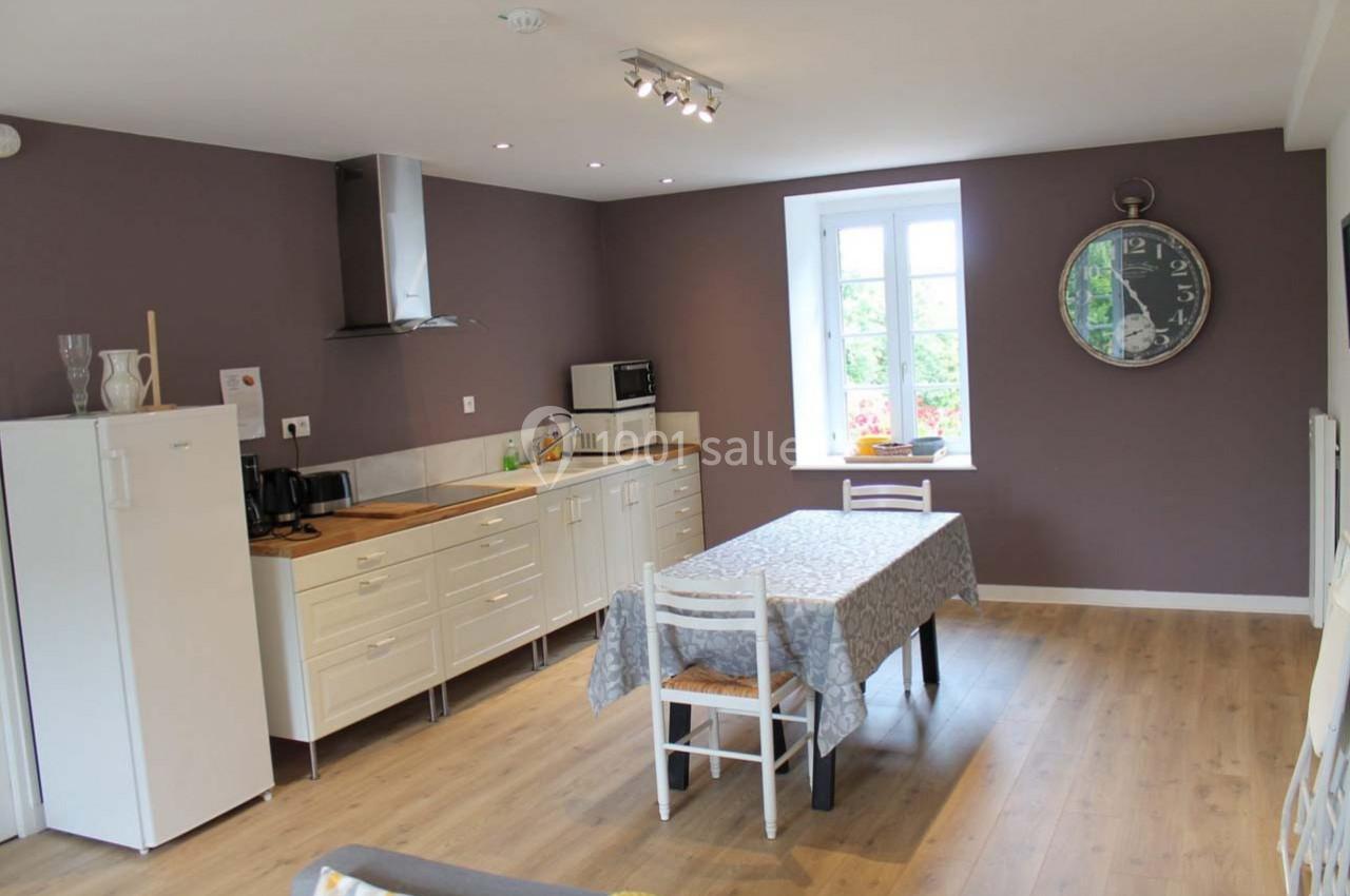 Cuisine lumineuse avec murs violets, meubles blancs, table avec nappe et grande horloge murale.