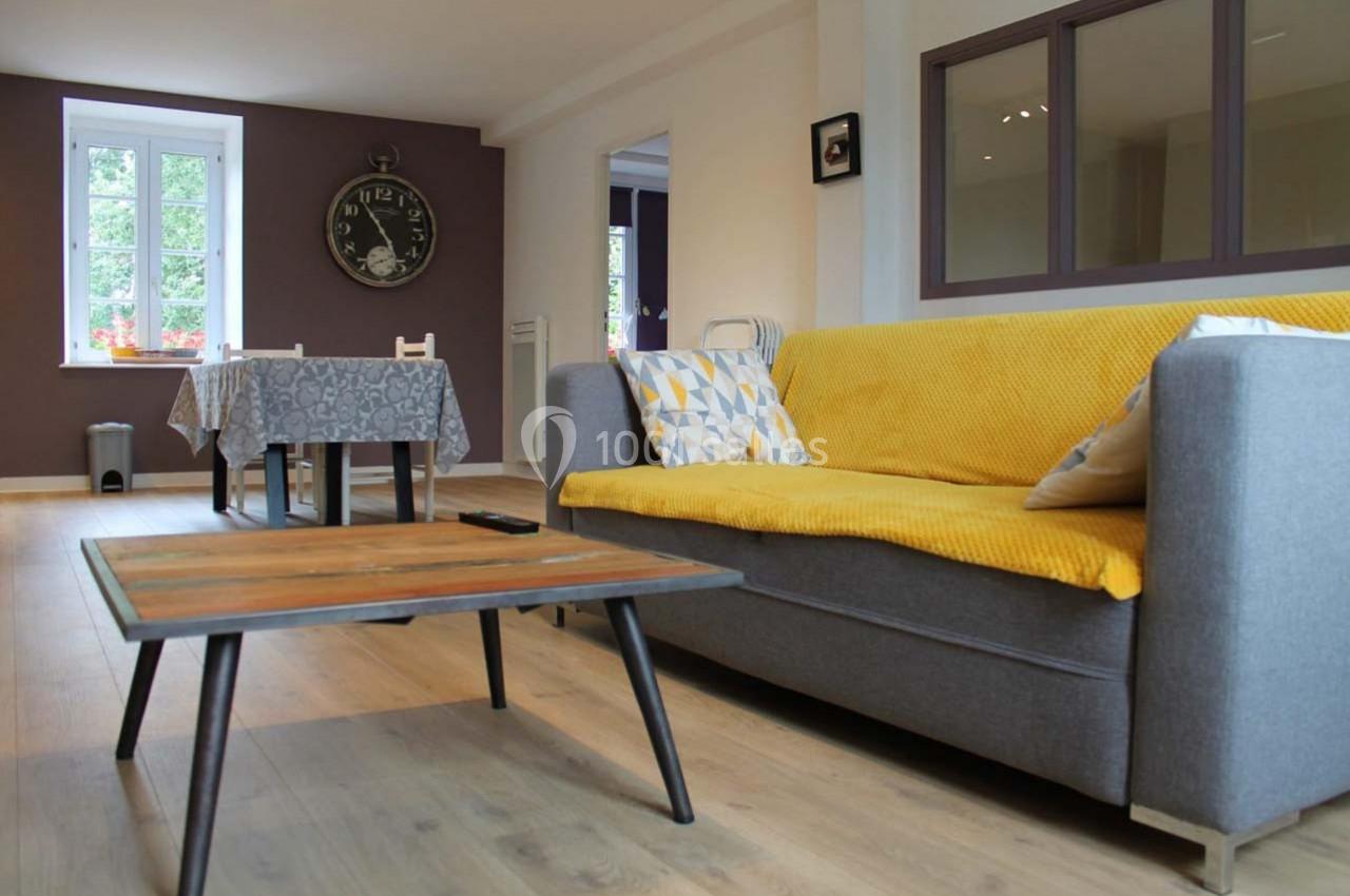 Salon avec canapé gris, plaid jaune, table basse en bois, et salle à manger en arrière-plan près d'une fenêtre.