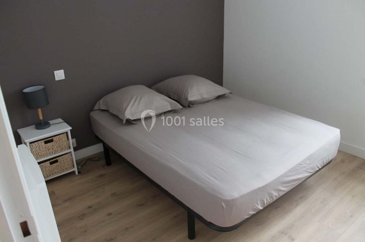 Lit double avec draps gris, deux oreillers assortis, table de chevet blanche avec lampe, sol en parquet clair.