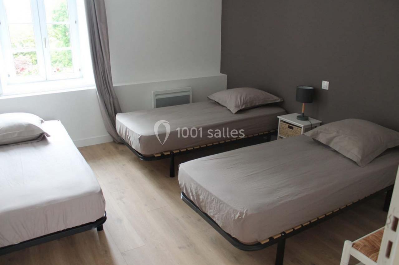 Trois lits simples avec draps gris dans une chambre lumineuse aux murs blancs et taupe, parquet clair au sol.