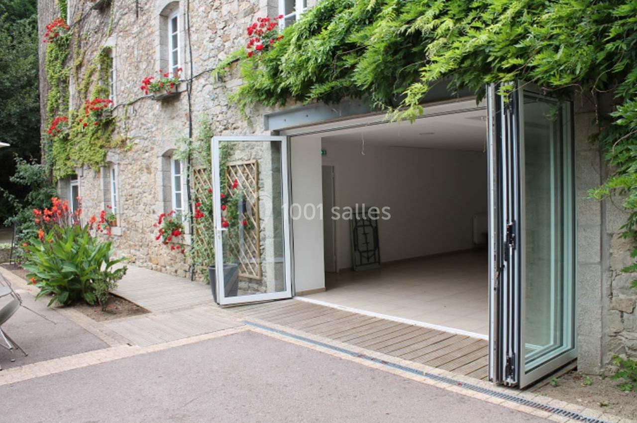 Façade en pierre avec porte vitrée pliable ouverte sur un intérieur vide, entourée de végétation et de fleurs rouges.