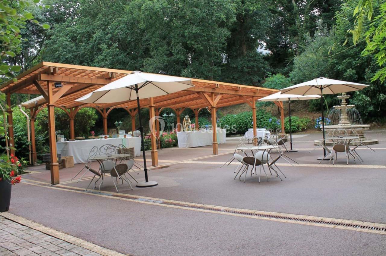 Terrasse extérieure avec pergolas en bois, tables et chaises en métal, parasols blancs et fontaine décorative.