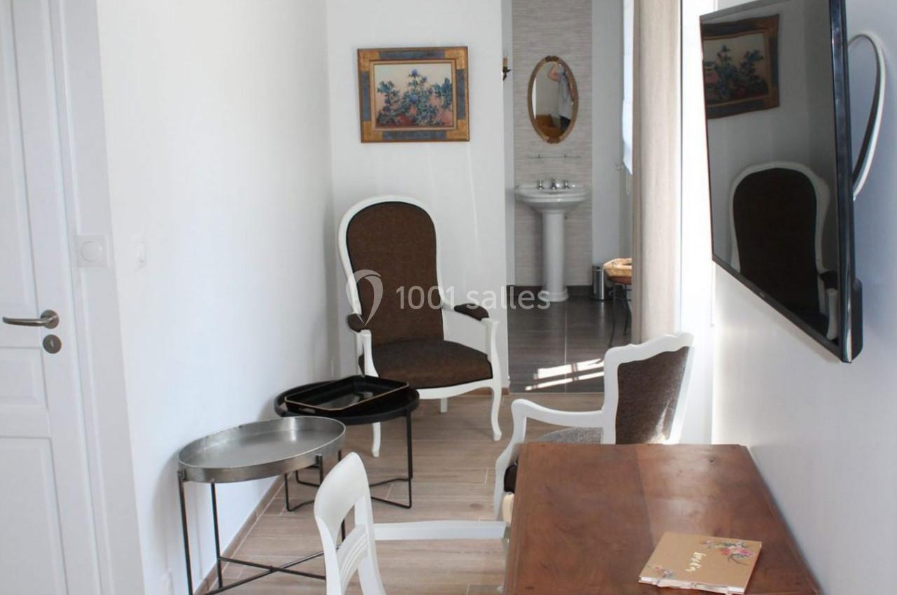 Salon lumineux avec fauteuils anciens, table en bois, télévision murale et vue sur un lavabo dans une pièce adjacente.