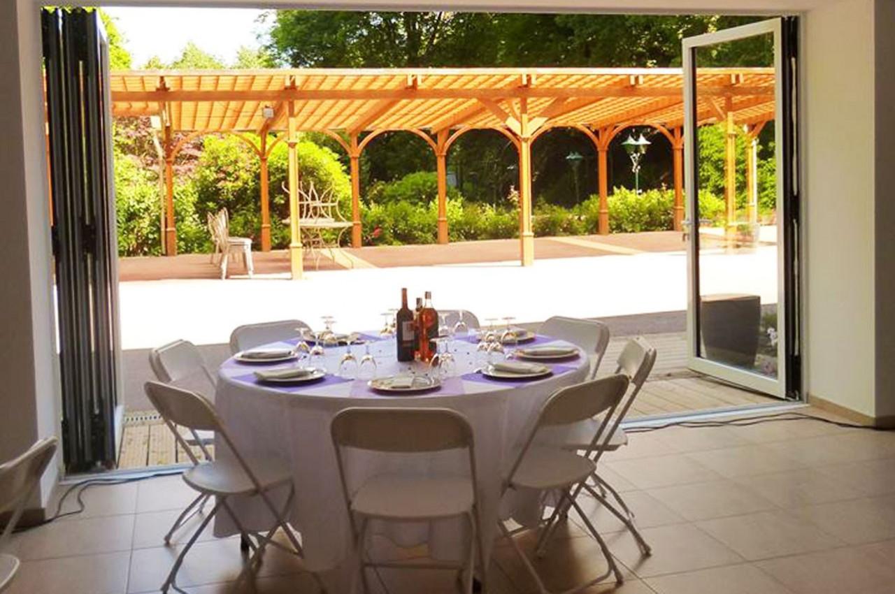 Table ronde dressée avec vaisselle et bouteilles, placée dans une salle ouverte sur une terrasse couverte en bois.