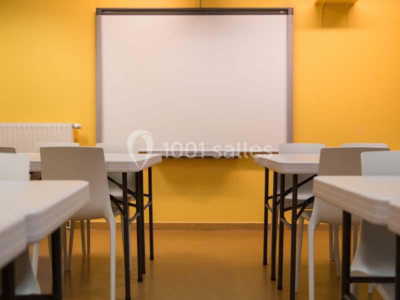 Salle de classe vide avec des tables blanches, des chaises et un tableau blanc sur un mur jaune.