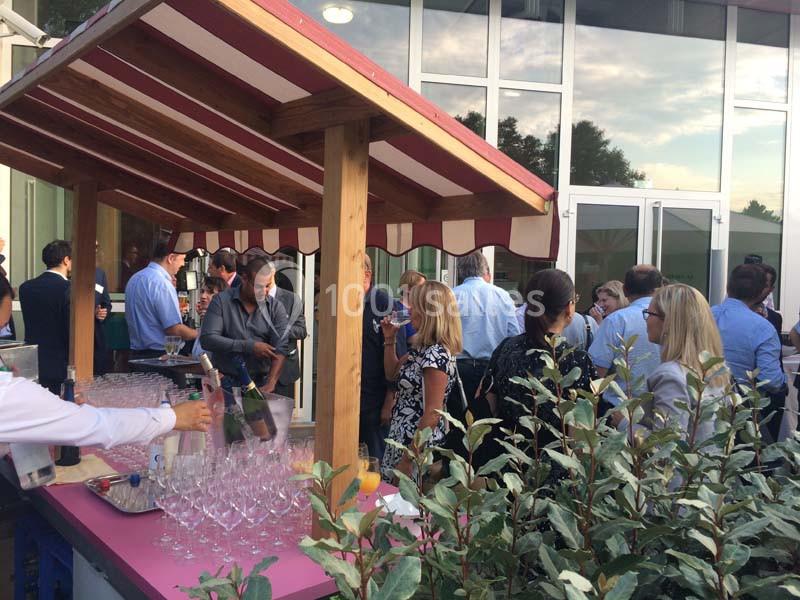 Un groupe de personnes échange autour d'un stand en bois avec des verres et des boissons, en extérieur près d'un bâtiment.