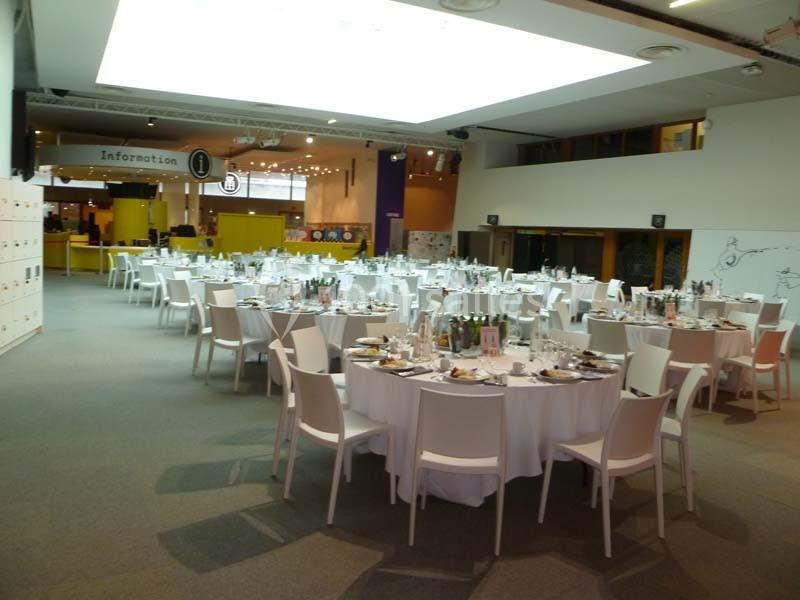 Salle de réception avec des tables rondes dressées pour un repas, entourées de chaises blanches, dans un espace lumineux.