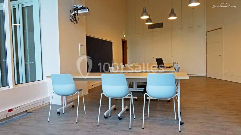 Salle de réunion moderne avec une longue table, des chaises bleues et un projecteur fixé au plafond.