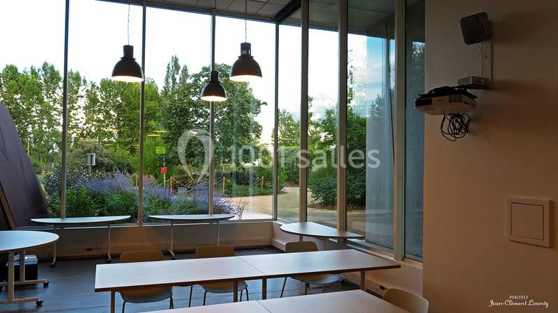 Salle lumineuse avec de grandes baies vitrées donnant sur un jardin arboré, tables et chaises disposées simplement.