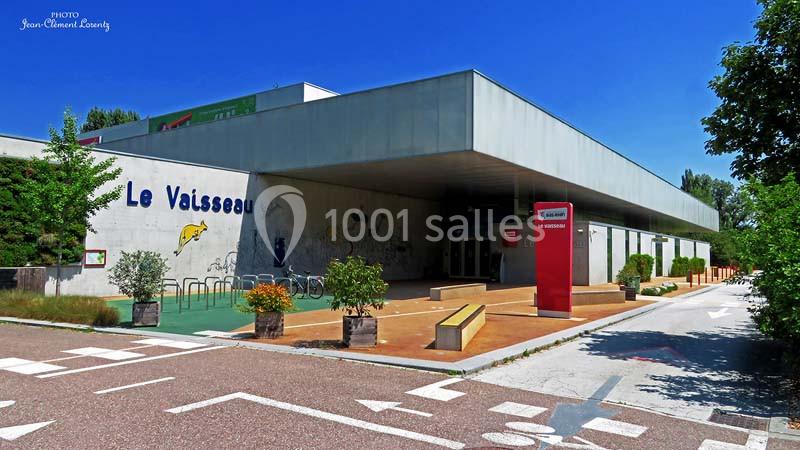 Façade moderne du bâtiment ’Le Vaisseau’ avec entrée principale, panneaux et espace extérieur aménagé.