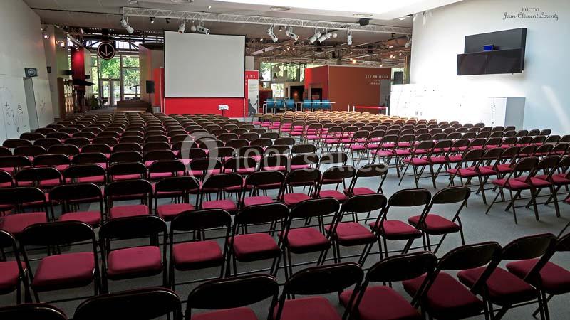 Salle de conférence avec de nombreuses rangées de chaises rouges vides, écran de projection et éclairage naturel.