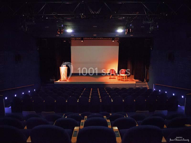 Salle de conférence vide avec un écran de projection, un pupitre et deux fauteuils éclairés sur scène.