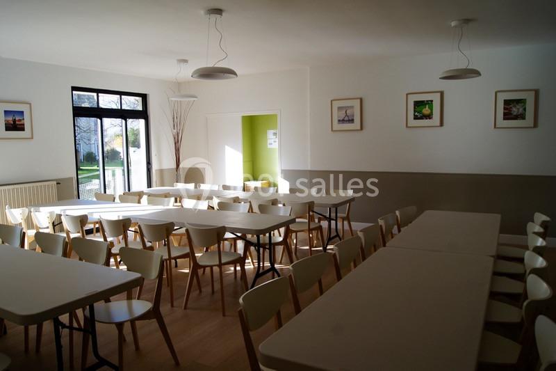 Salle lumineuse avec des tables et des chaises alignées, décorée de cadres au mur et éclairée par une grande baie vitrée.
