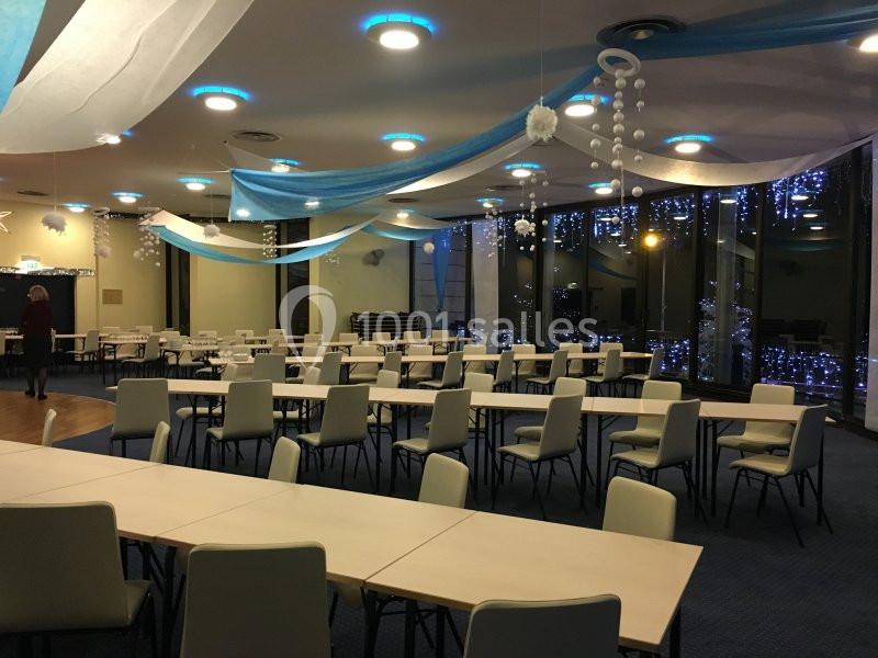 Salle de réception décorée avec des guirlandes lumineuses, des rubans bleus et blancs, tables et chaises alignées.