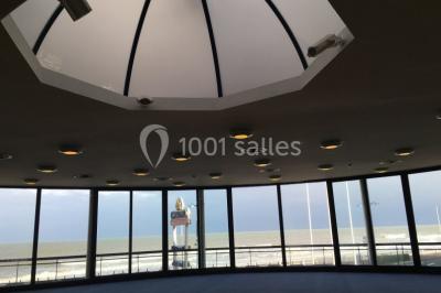 Vue intérieure d'une salle circulaire avec de grandes baies vitrées donnant sur la mer et un ciel partiellement nuageux.