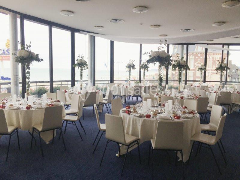 Salle de réception lumineuse avec tables rondes dressées, nappes blanches et décorations florales, vue sur l'extérieur.