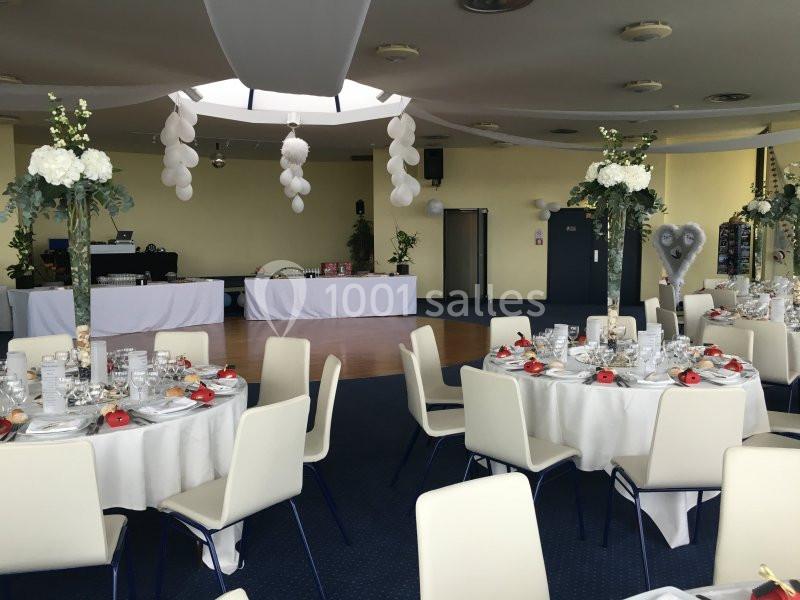 Salle de réception décorée avec des tables rondes dressées, des fleurs blanches et des ballons suspendus.
