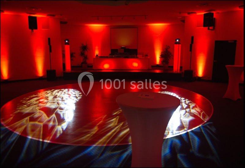 Salle de réception éclairée en rouge avec une piste de danse illuminée et des tables hautes en premier plan.