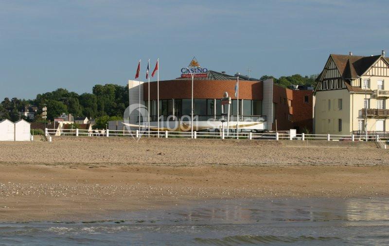 Location salle Villers-sur-Mer (Calvados) - Salle Panoramique Villers Sur Mer #6 Bâtiment moderne d'un casino en bord de plage, entouré de maisons et de verdure, avec des drapeaux sur le toit.