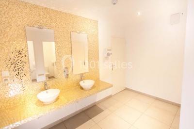 Salle de bain lumineuse avec deux lavabos, miroirs et murs ornés de mosaïques dorées.