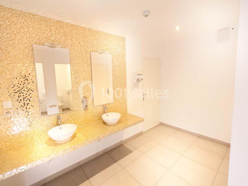 Salle de bain lumineuse avec deux lavabos, miroirs et murs ornés de mosaïques dorées.