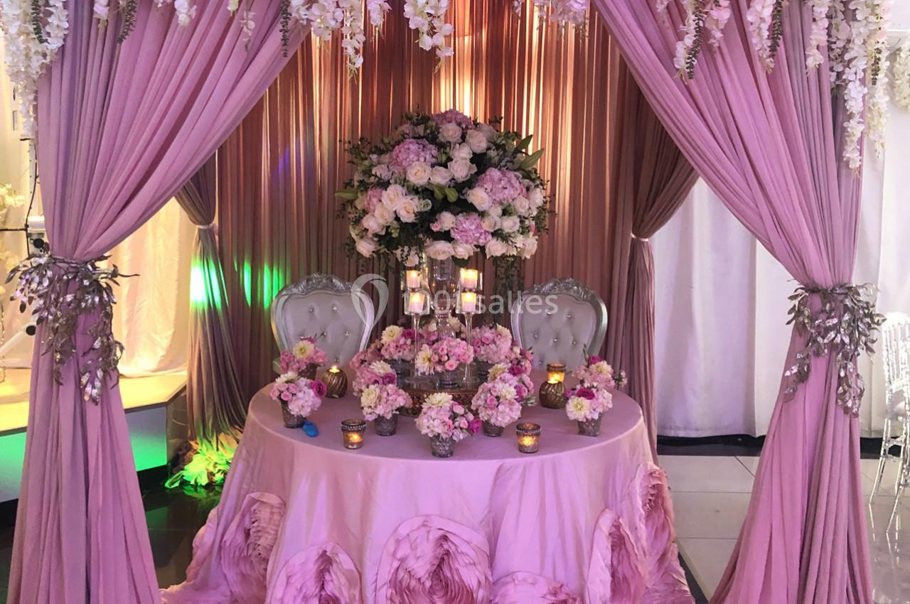 Table décorée pour un mariage avec nappes roses, chaises élégantes, fleurs et bougies sous un dais rose.