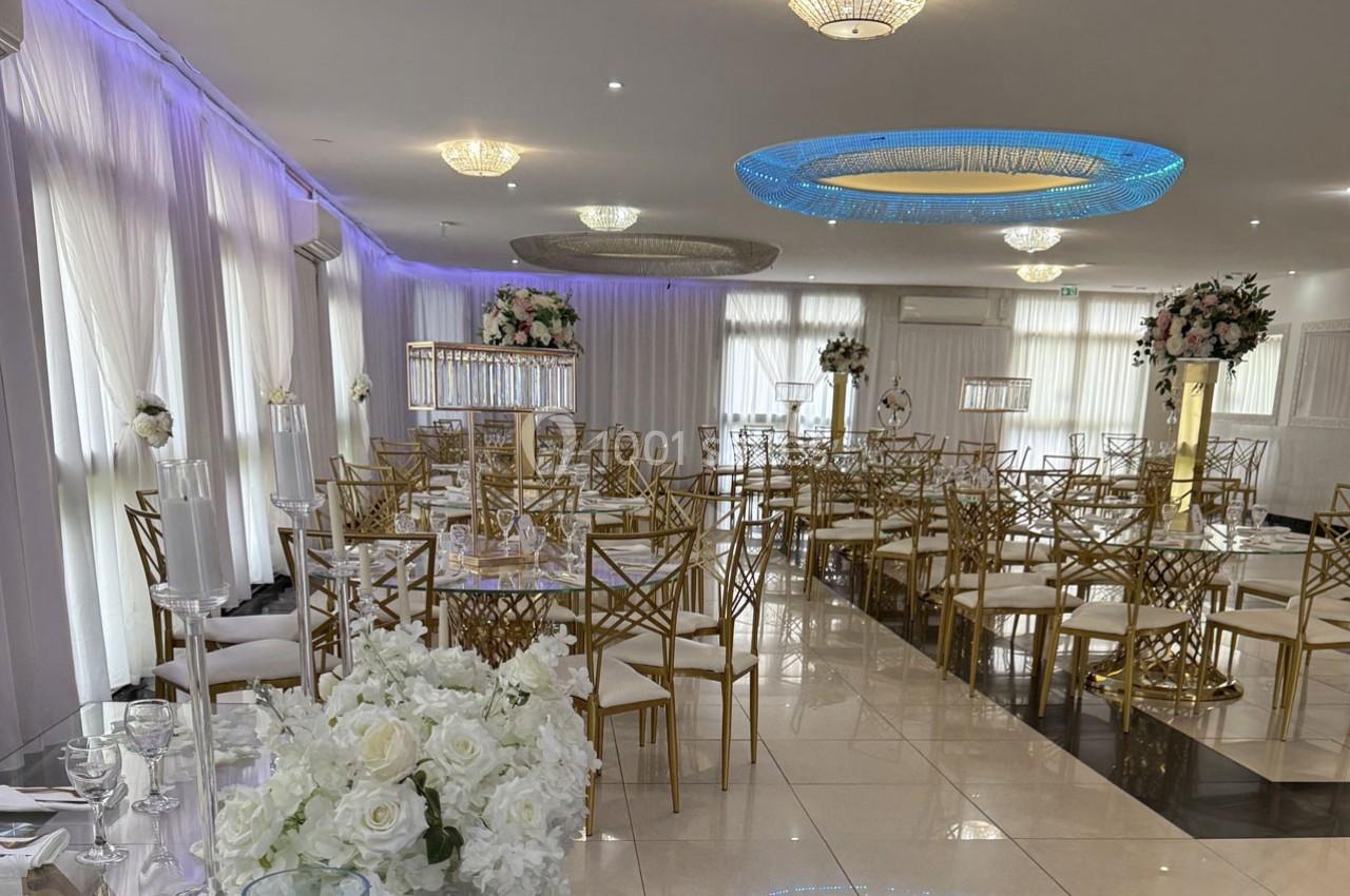 Salle de réception élégante avec tables décorées, chaises dorées et arrangements floraux blancs sous un éclairage moderne.