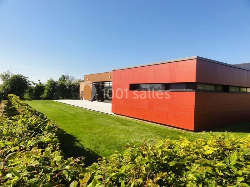 Bâtiment moderne avec façade rouge, large terrasse et pelouse entourée de haies sous un ciel dégagé.