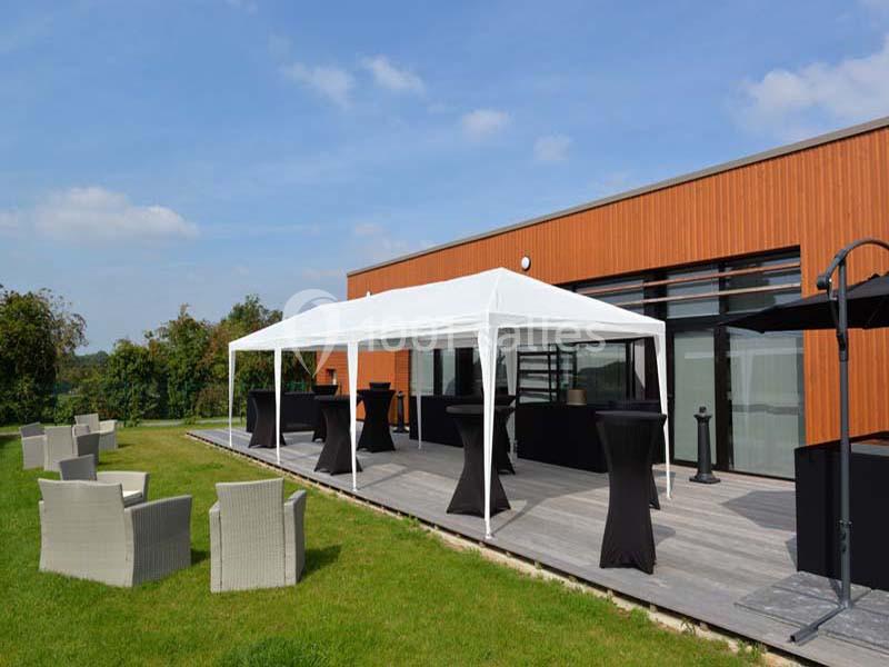 Tente blanche installée sur une terrasse en bois avec tables hautes noires, entourée de chaises et d'un jardin.