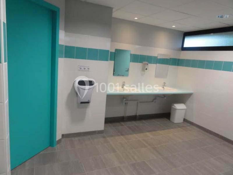 Salle de bain publique avec deux lavabos, un miroir, une porte turquoise et un distributeur de papier mural.