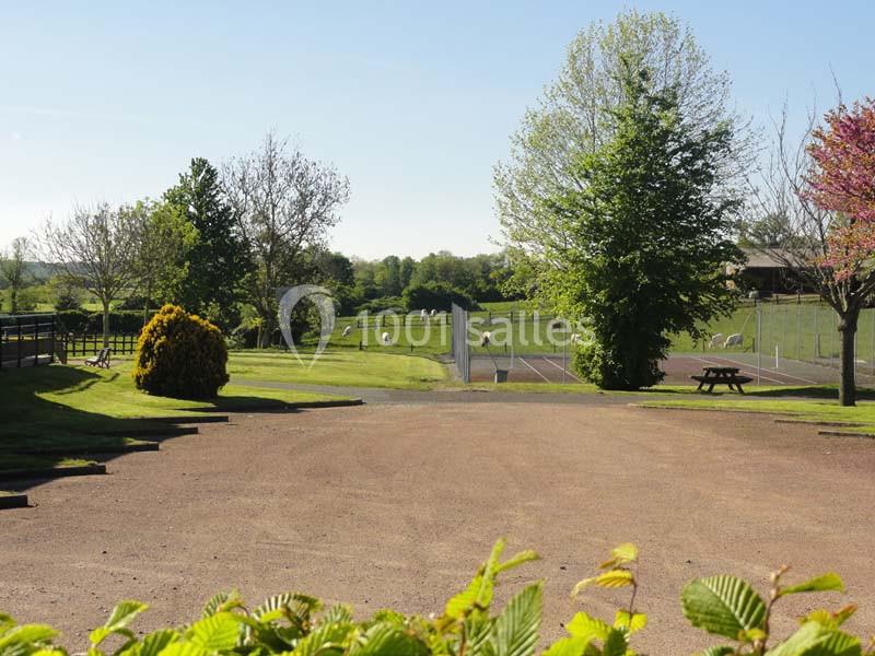 Allée en gravier bordée de verdure, menant à des terrains de sport et des pâturages sous un ciel dégagé.
