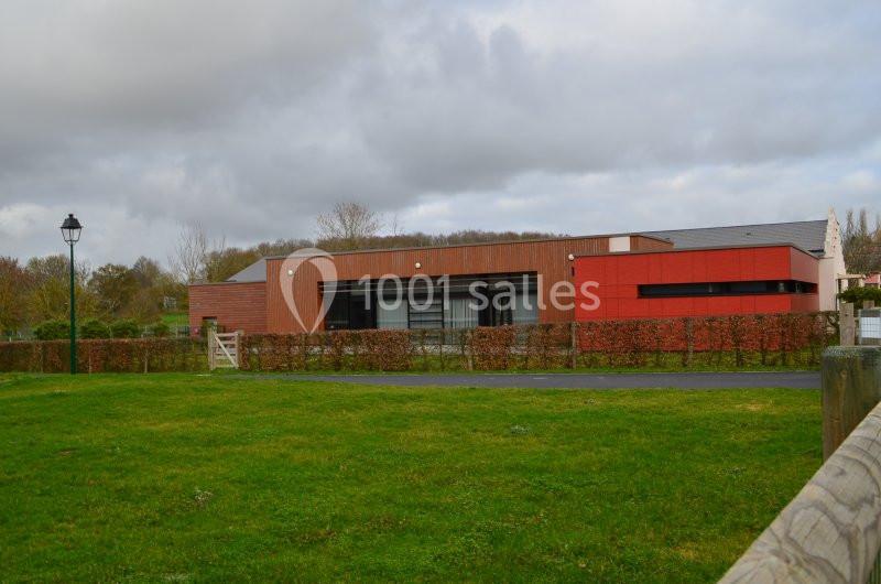 Bâtiment moderne en bois et rouge entouré de haies, avec pelouse et lampadaire au premier plan sous un ciel nuageux.