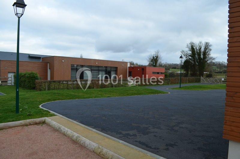 Bâtiment moderne en bois et briques entouré d'une haie, avec un parking asphalté et un lampadaire au premier plan.