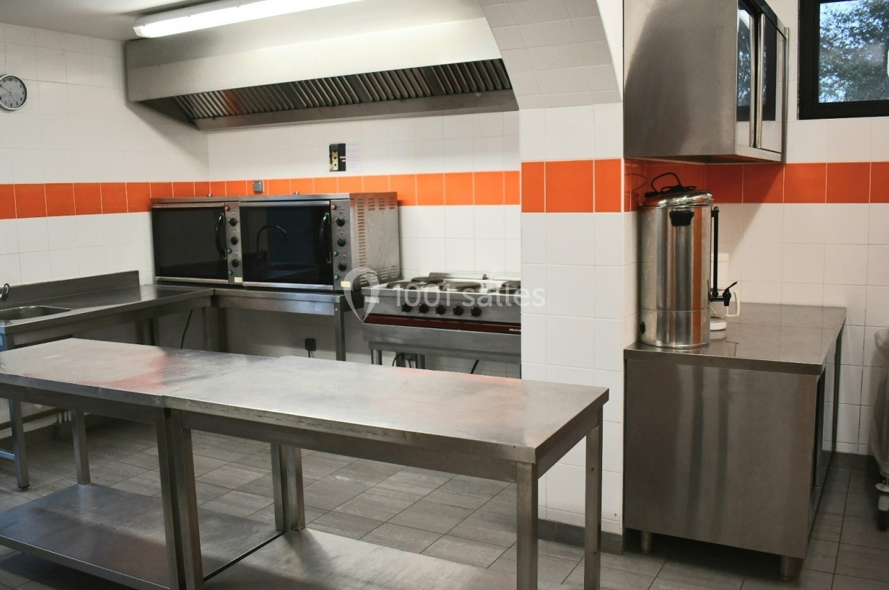 Cuisine professionnelle équipée avec plans de travail en inox, four, plaques de cuisson et carrelage mural blanc et orange.
