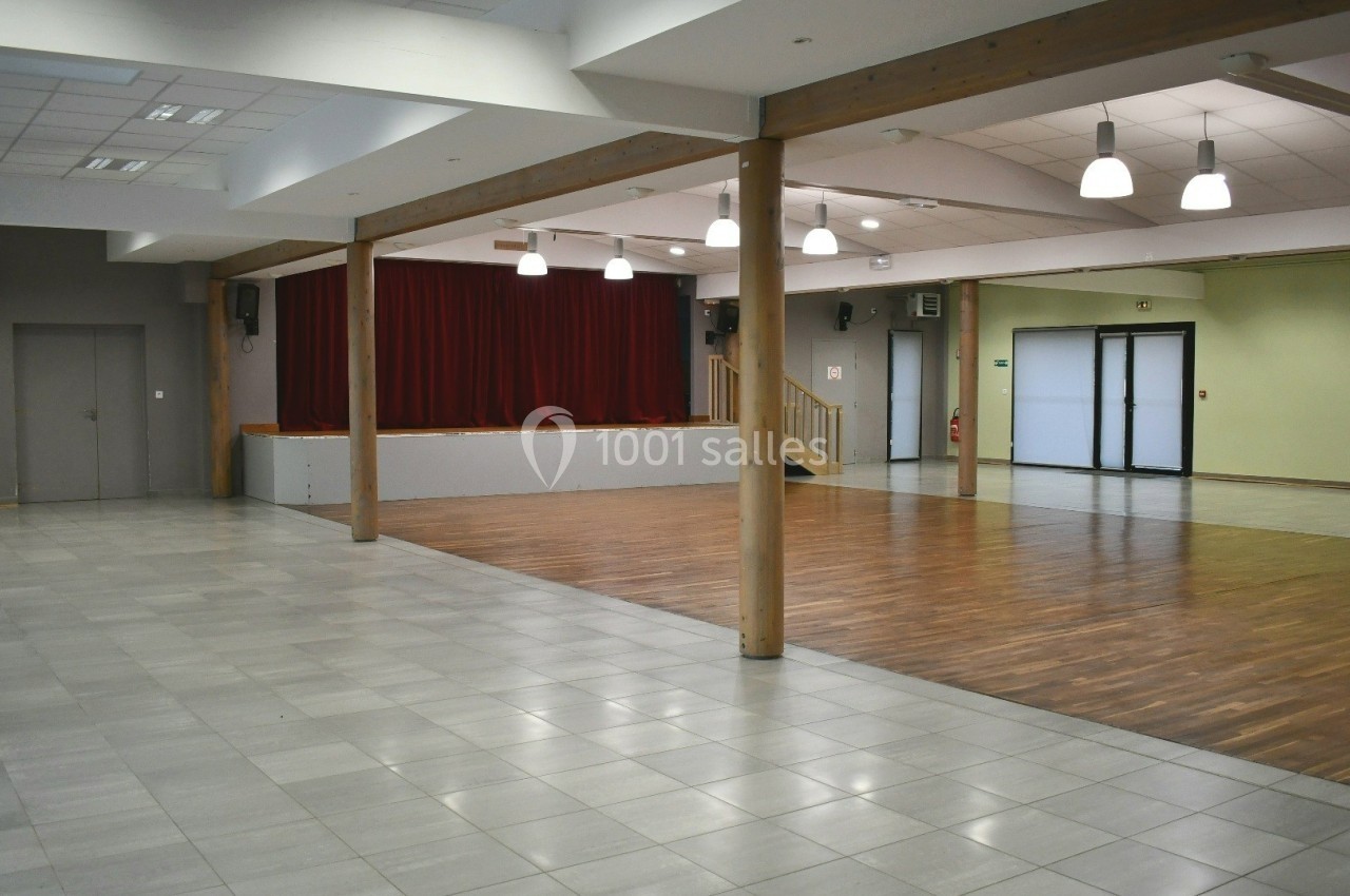 Salle polyvalente vide avec sol en carrelage et parquet, scène avec rideaux rouges et éclairage suspendu.