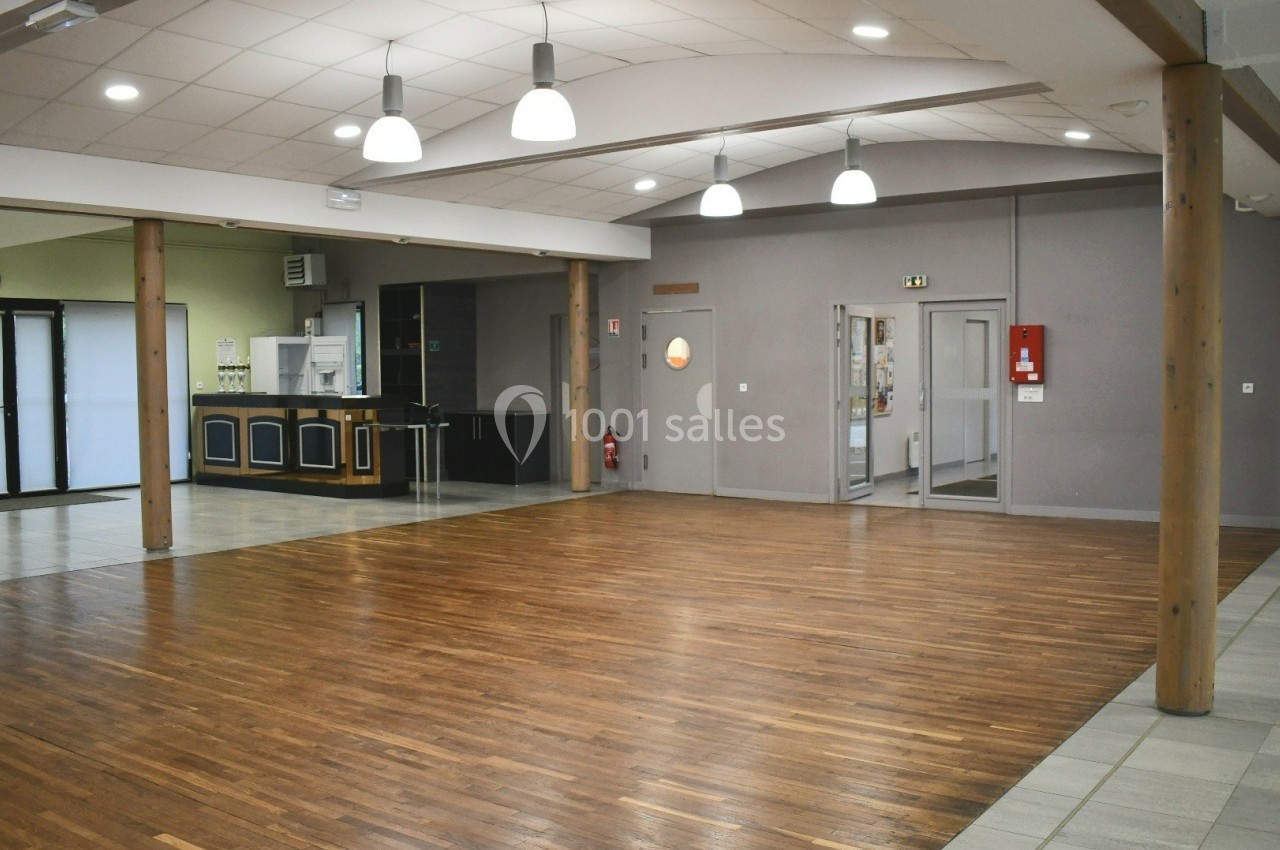 Salle polyvalente vide avec parquet, murs clairs, éclairage suspendu et coin cuisine en arrière-plan.