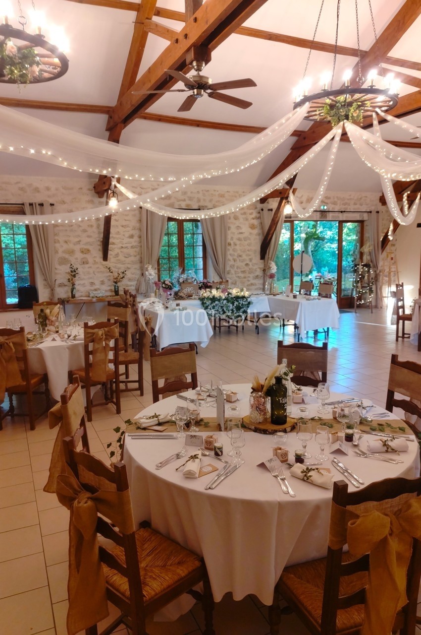 Salle de réception décorée pour un mariage avec tables rondes dressées, guirlandes lumineuses et voilages suspendus.