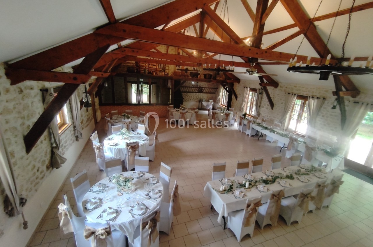 Salle de réception lumineuse avec poutres apparentes, tables décorées pour un événement et grandes fenêtres.